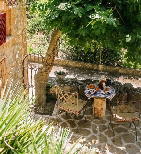 Breakfast set on a rustic stone table under a fig tree in the walled courtyard, an old iron gate and dappled light.