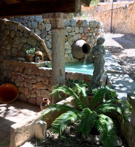 A dry-stone water feature with a spouting spring filling a small turquoise plunge basin, a stone figure and a palm beside it.