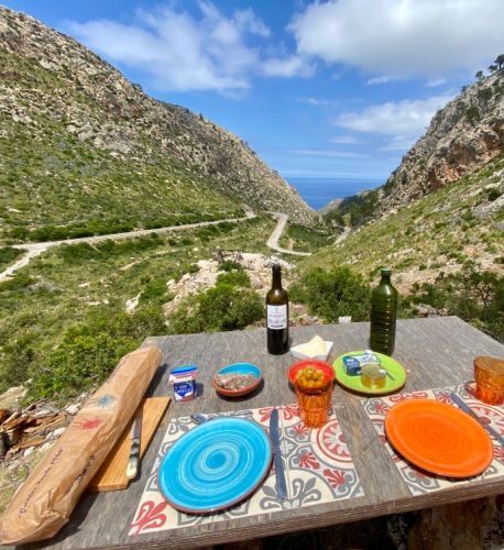 A table laid with bread and wine on the terrace edge, looking down the dry valley to a thin band of open sea between two mountains.
